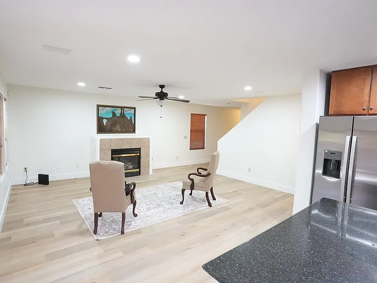 Open living area with recessed lighting, ceiling fan, and a fireplace, flowing into the kitchen with stainless fridge.