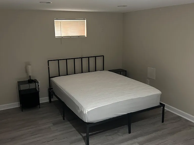 Furnished bedroom with a metal bed frame, nightstands, and durable wood-look flooring, plus a window with blinds.