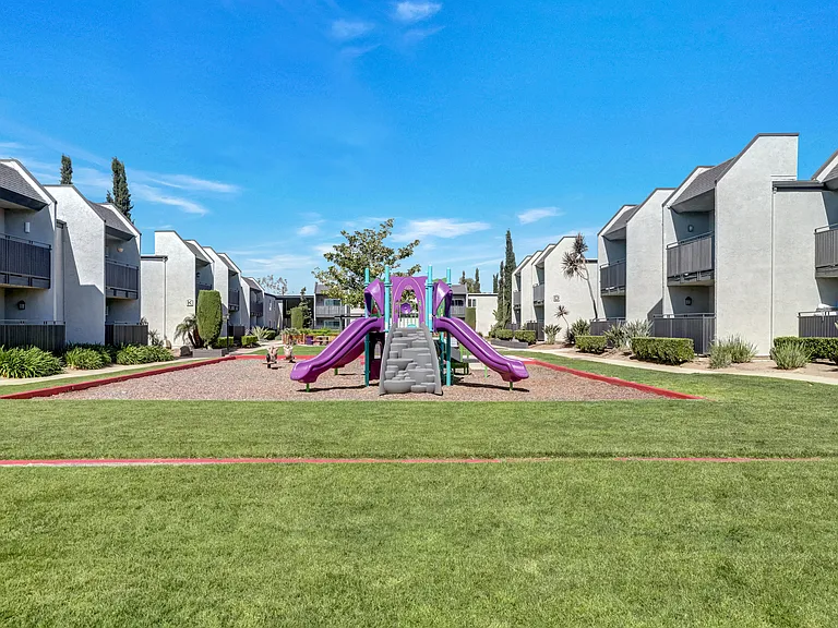 A playground with a purple slide in the middle of a grassy area with apartments in the background.