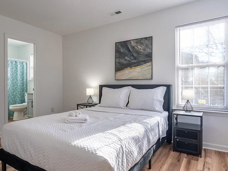 Furnished bedroom with a queen bed, nightstands, and a large window with blinds; adjacent bath access for convenience.