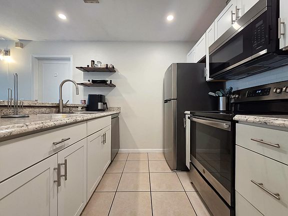 Updated galley kitchen with shaker cabinets, stone-look counters, tile floors, and stainless steel appliances.