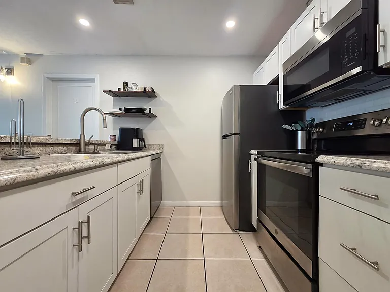 Updated galley kitchen with shaker cabinets, stone-look counters, tile floors, and stainless steel appliances.