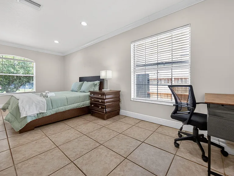 Spacious bedroom with tile flooring, large windows, and a dedicated desk area alongside a furnished sleeping space.