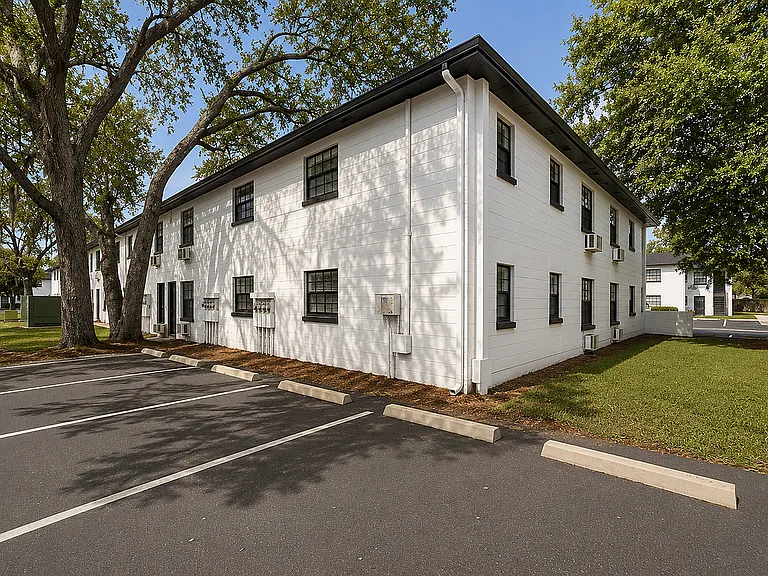 Charming white two-story apartment building with lush greenery, spacious parking, and serene surroundings.