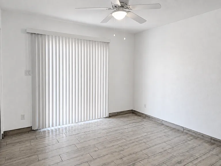 Spacious primary bedroom featuring continuous wood-look tile flooring, fresh neutral paint, and a modern ceiling fan. The large sliding glass door opens directly to your private, fully fenced yard.