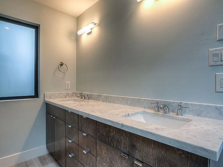 Master bath dual vanity from the opposite angle _ Carrara marble counter, dark-stained cabinetry, sconce lighting, and a black-framed picture window