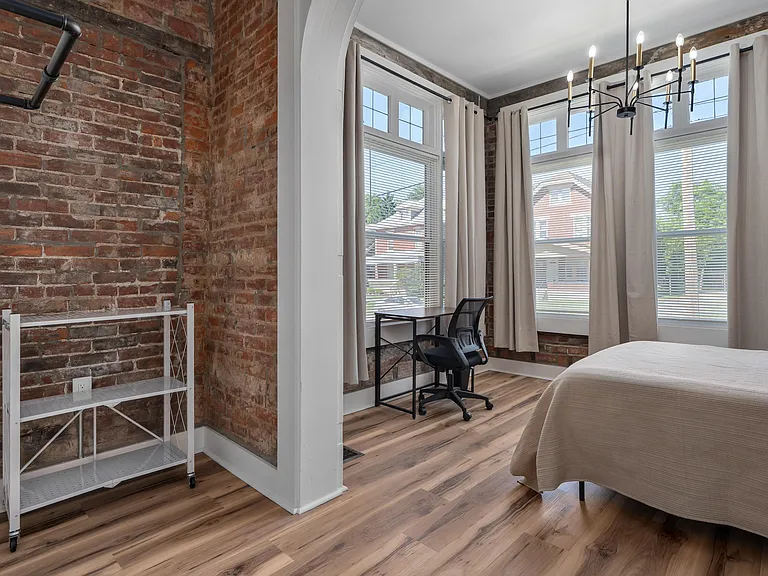 Bedroom features exposed brick accents, large windows with curtains, and space for a bed plus a dedicated work desk.