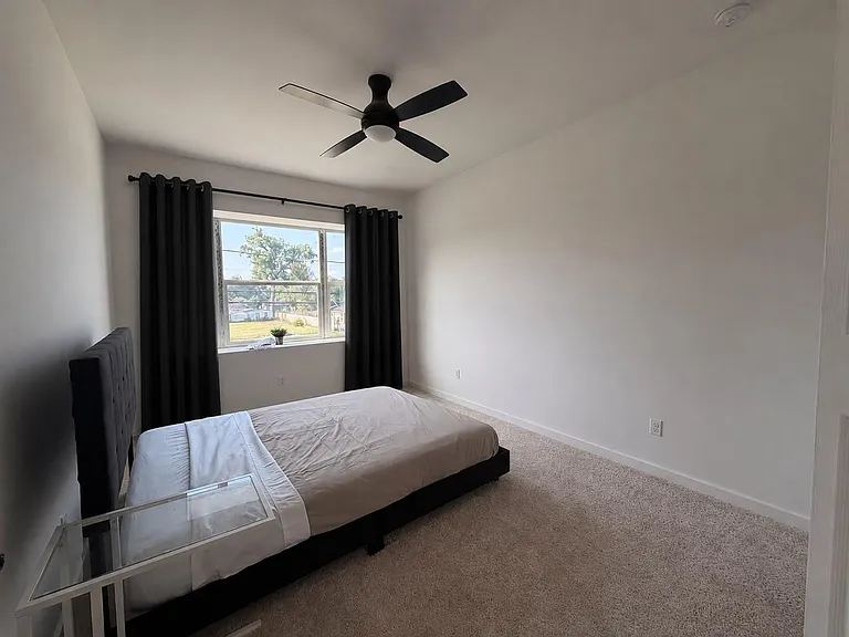 Spacious bedroom with a large window and blackout curtains, plush carpet, and a ceiling fan for year-round comfort.