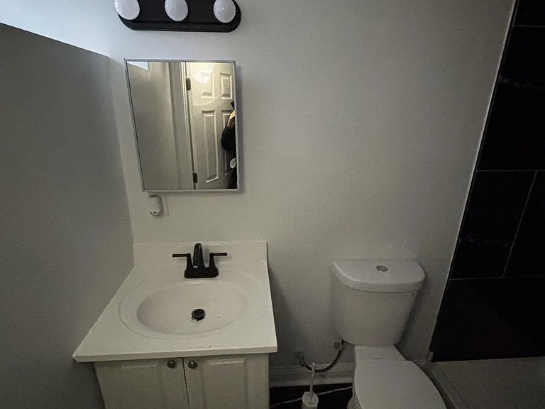 Compact bathroom with a single-sink vanity, mirror lighting, and a full tub/shower surround with dark tile flooring.