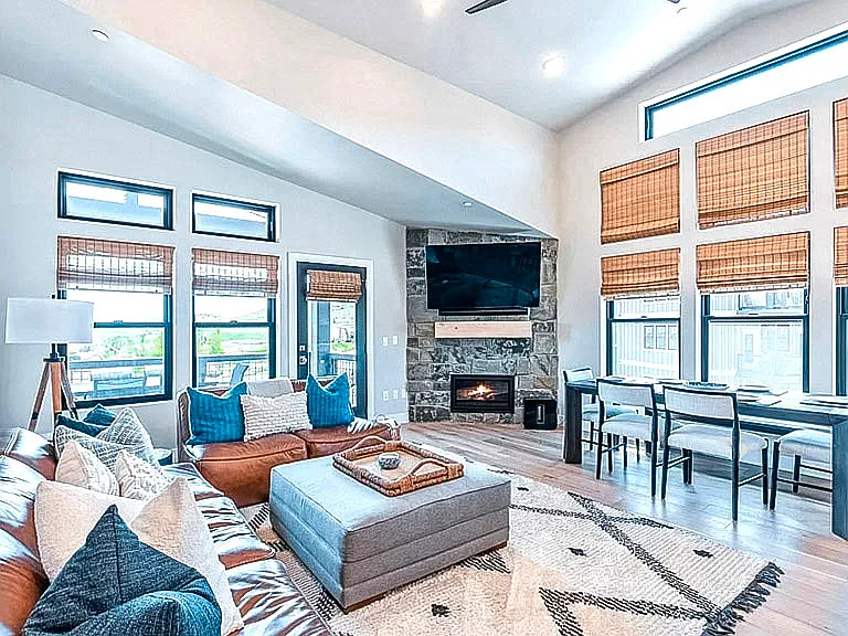 Bright living room with stone fireplace, wall-mounted TV, soaring ceilings, and bamboo blinds on large windows