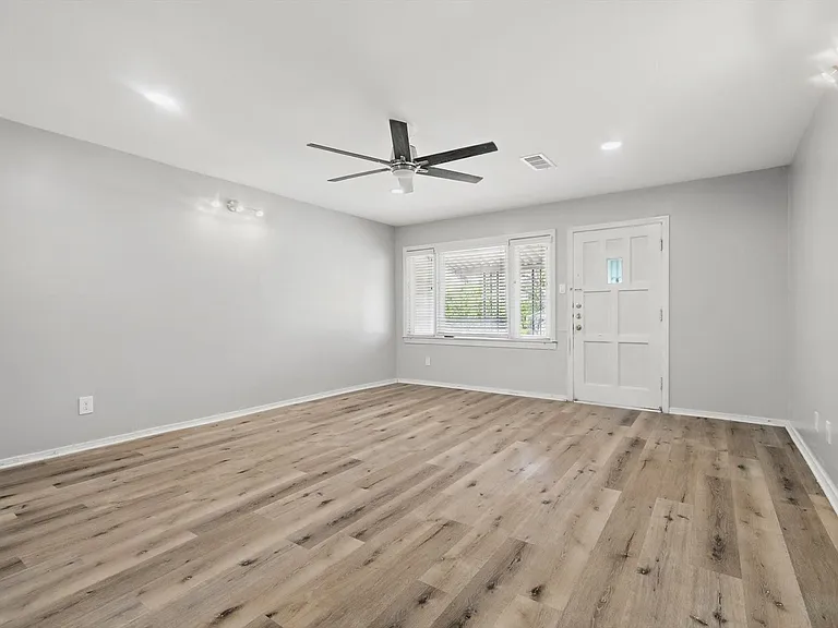 Spacious living room with neutral soft grey paint, LVP flooring, recessing lighting, a ceiling fan and ample amount of light from the large window.