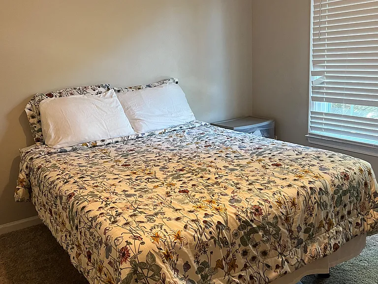 Furnished bedroom with a comfortable bed, carpeted floors, and a large window with blinds for controlled natural light.