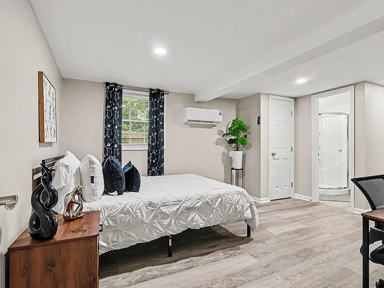 Furnished bedroom with a full bed, desk area, and a private en-suite bath, plus a window with curtains and mini-split AC.
