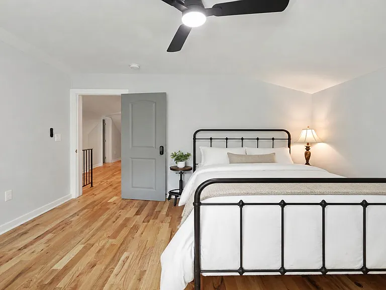 Spacious bedroom with hardwood floors, ceiling fan, and a comfortable bed setup with bedside lighting and easy hallway access