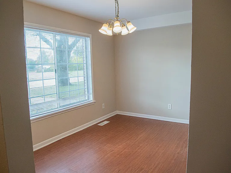 Bright, inviting room with large window and elegant light fixture, featuring hardwood floor and soft beige walls, perfect for a cozy dining or living area.
