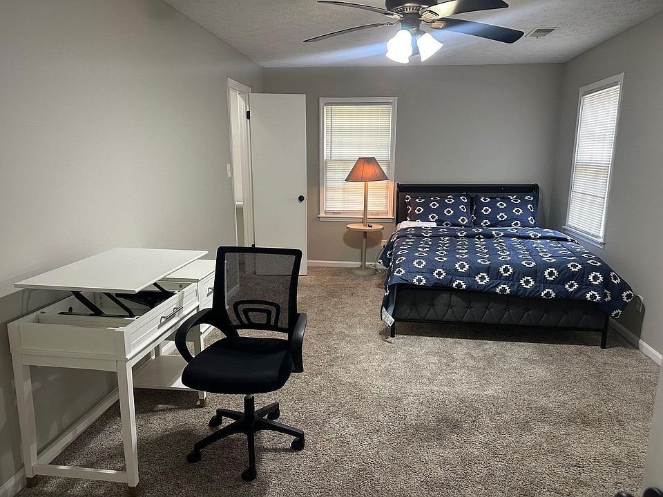 Furnished bedroom with a full-size bed, dedicated desk workspace, and ceiling fan, plus two windows with blinds.