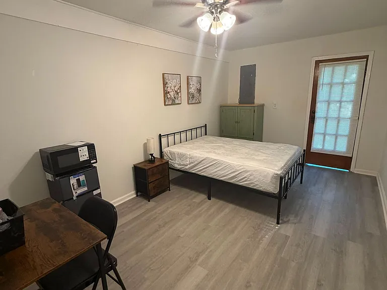 Furnished bedroom with bed frame, nightstand, dresser, and a small desk area, plus ceiling fan and door with blinds.