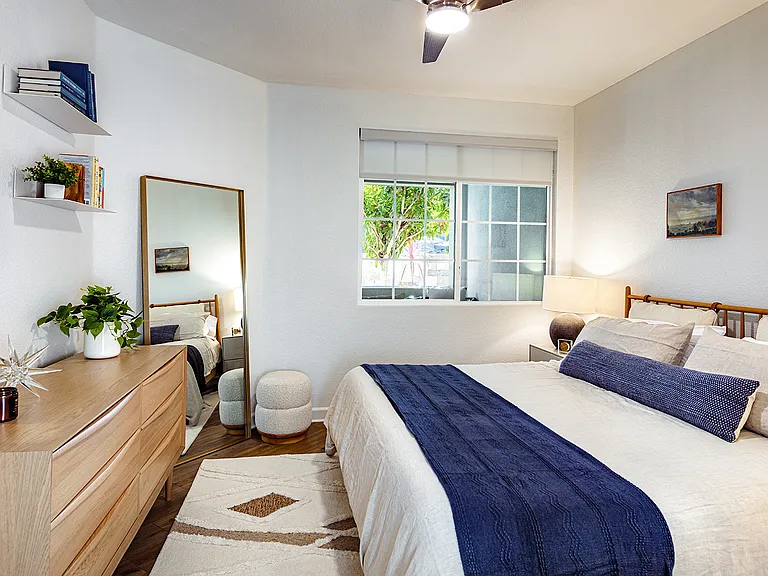 A bedroom with a dresser, a floor-length mirror, a large window, and a bed with a blue throw.