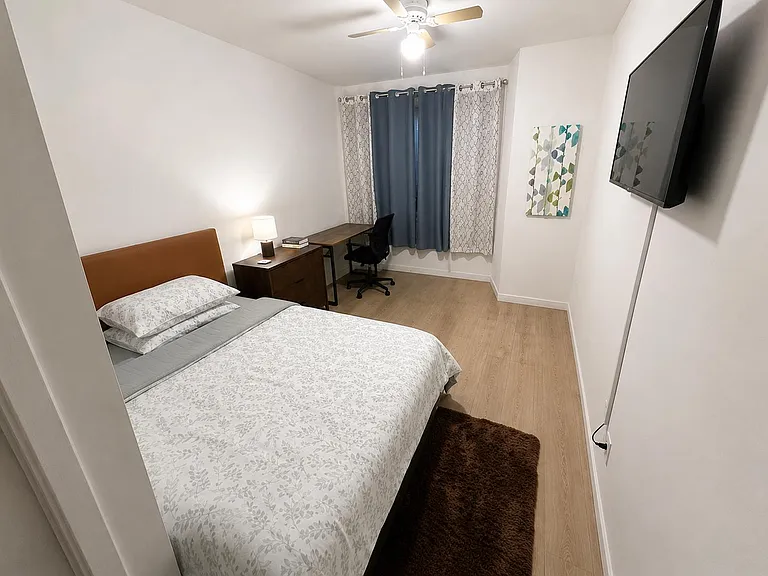 Comfortable furnished private bedroom with a queen bed, TV, ceiling fan, window, and a full private bathroom.