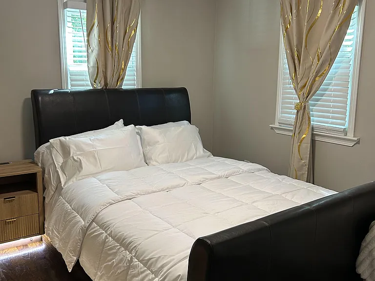 Furnished bedroom with a queen bed, nightstand, and two windows with blinds and curtains for adjustable light and privacy.