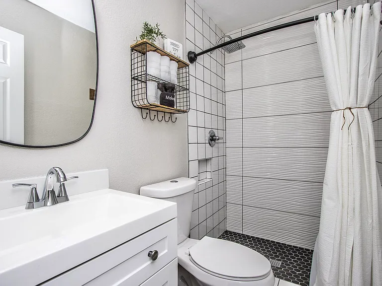 Modern bathroom shower at Casa Del Sol, Tempe rental home. Features sleek white grid and wavy textured tile, a luxury rain showerhead, a handheld sprayer, industrial black pipe hooks, and an inset storage niche. Premium amenity for a Tempe, AZ stay.