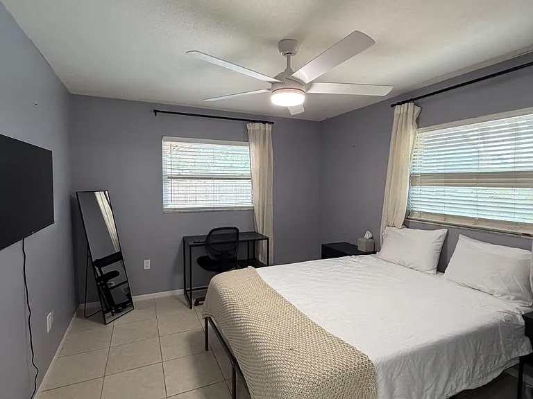 Private bedroom with tiled floors, ceiling fan, and two windows with blinds and curtains plus a desk workspace.