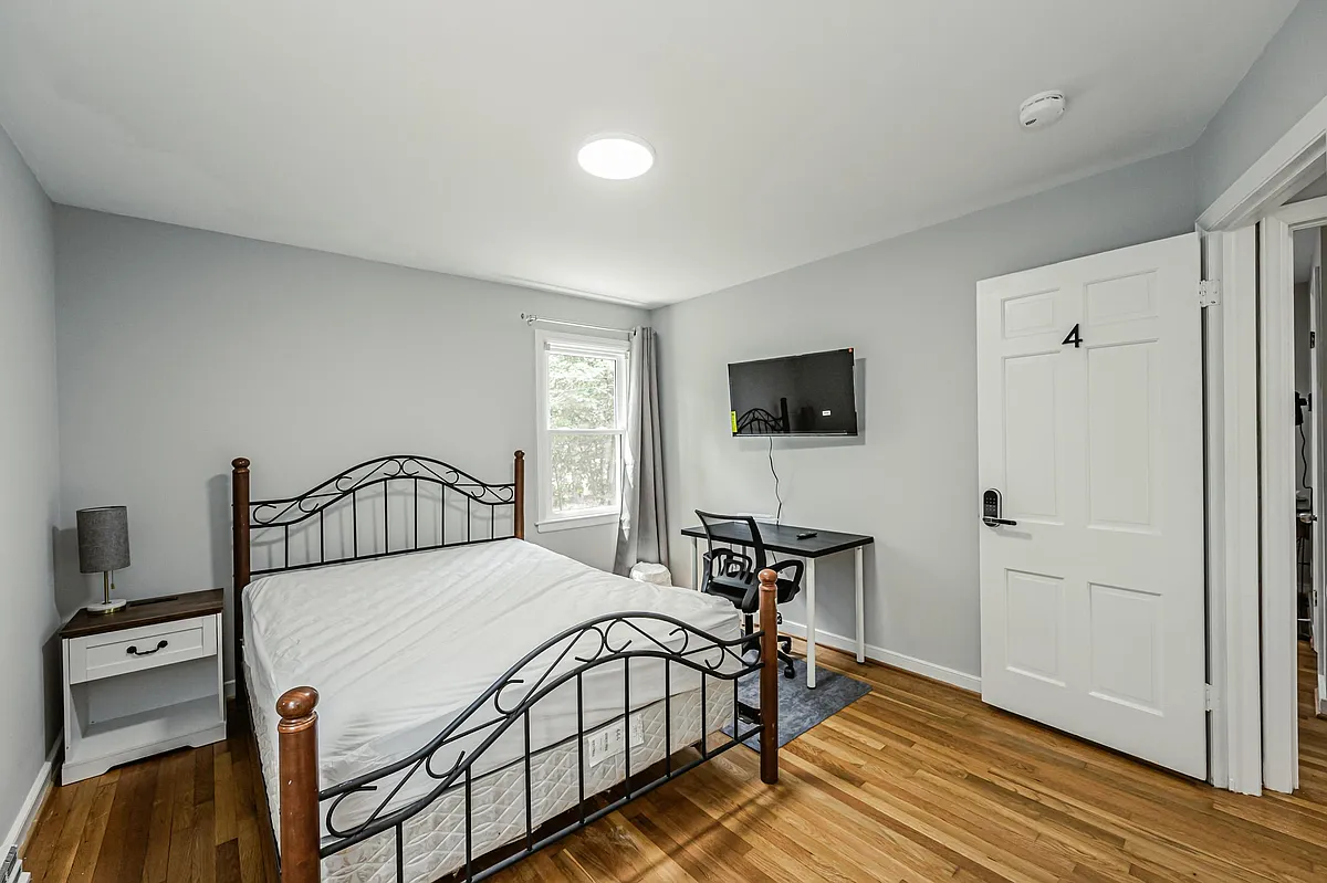 Private bedroom with hardwood floors, a furnished bed, work desk, and wall-mounted TV plus a window with curtains. - Stratford Ct