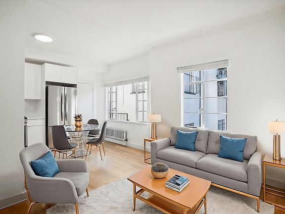 Living room with large window allowing natural light to flood the space, features a sofa. coffee table, dining table with chairs and a refrigerator