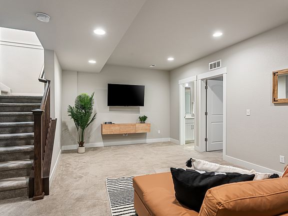 Cozy living room with carpeted floor, modern neutral walls, recessed lighting, and a comfortable sofa. Features a wall-mounted TV, floating wooden shelf, indoor plant, and a staircase with dark wooden railing. Bright, spacious and inviting atmosphere