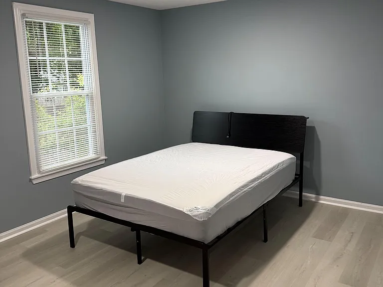 Furnished bedroom with a large window and blinds, durable plank-style flooring, and ample space for a nightstand or desk.