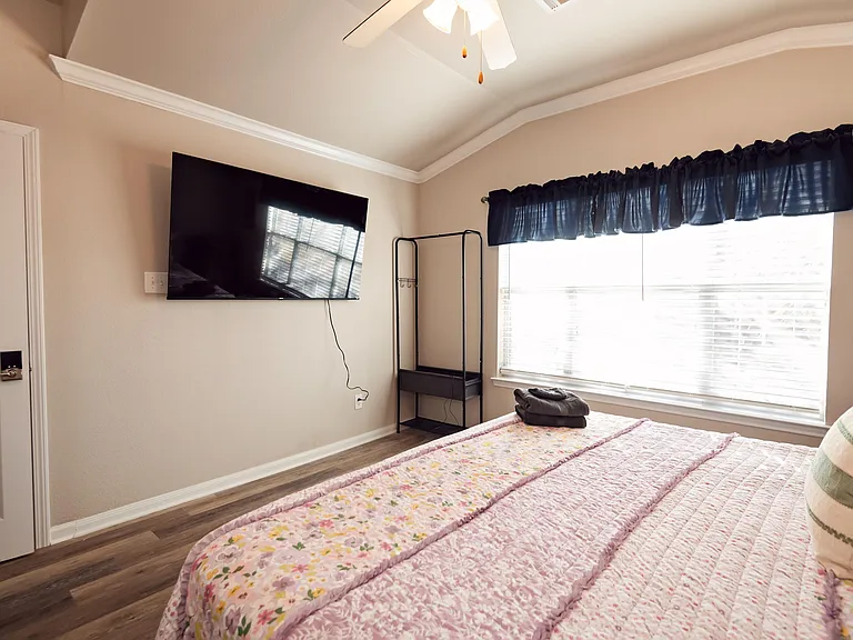 Spacious bedroom with a large window, ceiling fan, wall-mounted TV, and wood-style flooring for easy maintenance.