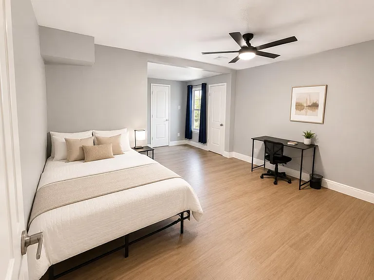 Spacious bedroom with a queen bed, dedicated desk area, ceiling fan, and window with curtains for natural light.