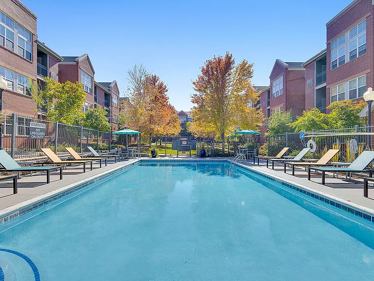 Inviting apartment complex with a sparkling blue swimming pool, ample lounge chairs, and vibrant autumn trees, surrounded by modern brick buildings.