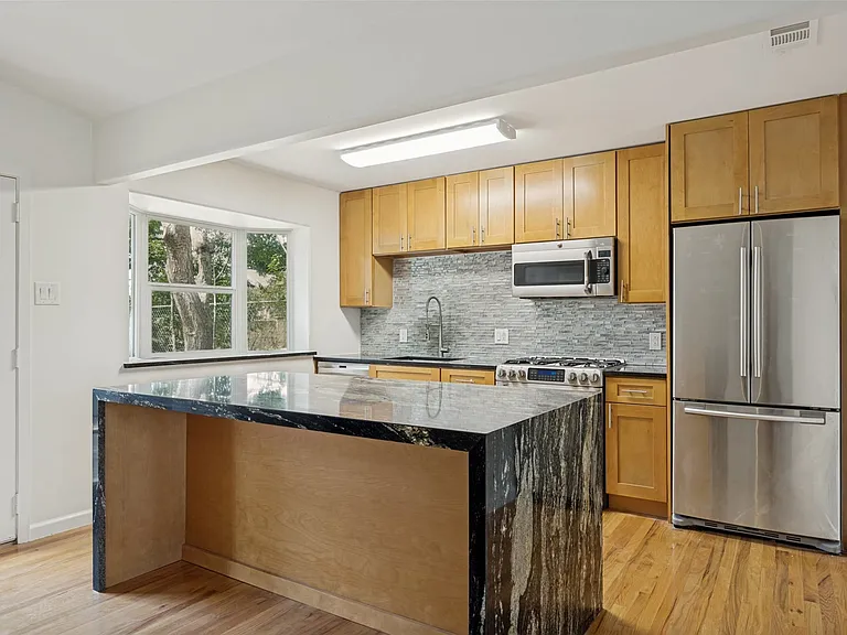 Kitchen featuring a large waterfall island with a granite countertop, stainless steel appliances, wood-finish flooring, light wood cabinetry, and a subway tile backsplash