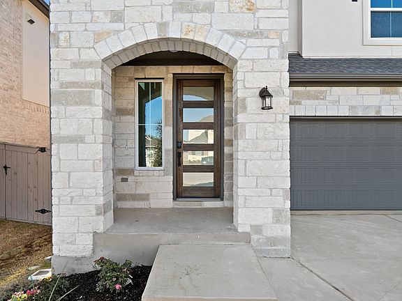 The image shows a modern, stone-clad house with an arched entryway, a wooden front door, and a garag
