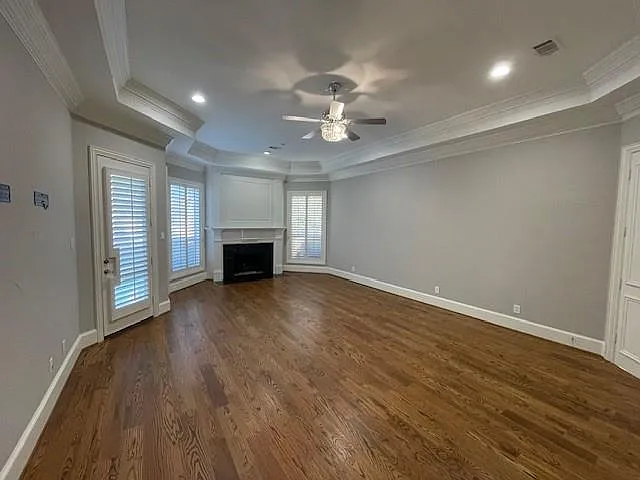 Unfurnished living room with a fireplace, a tray ceiling, a ceiling fan, dark wood-style flooring, and ornamental molding