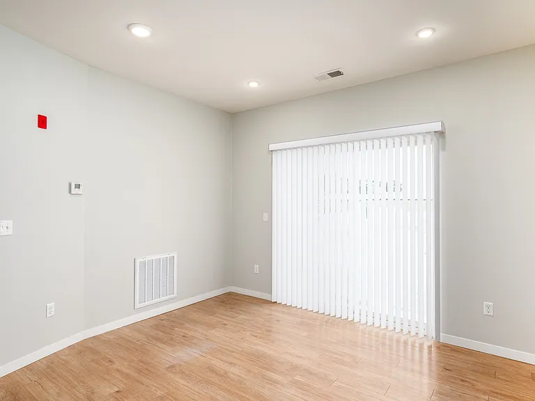 Bright and cozy room with sleek hardwood flooring, soft neutral walls, and large sliding glass doors with vertical blinds for natural light. Bright and cozy room with sleek hardwood flooring, soft neutral walls, and large sliding glass doors with vertical