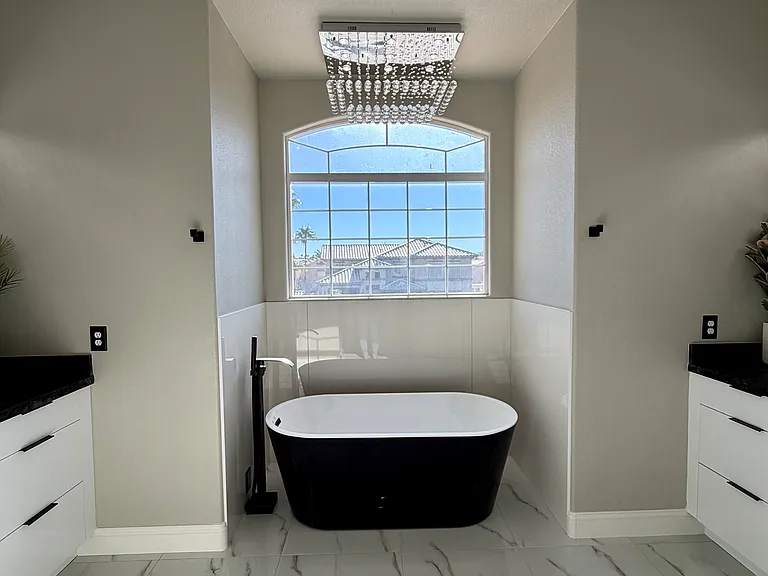 Luxurious primary bathroom featuring a freestanding soaking tub, dual vanities, glossy white cabinetry, granite countertops, and beautiful lighting.
