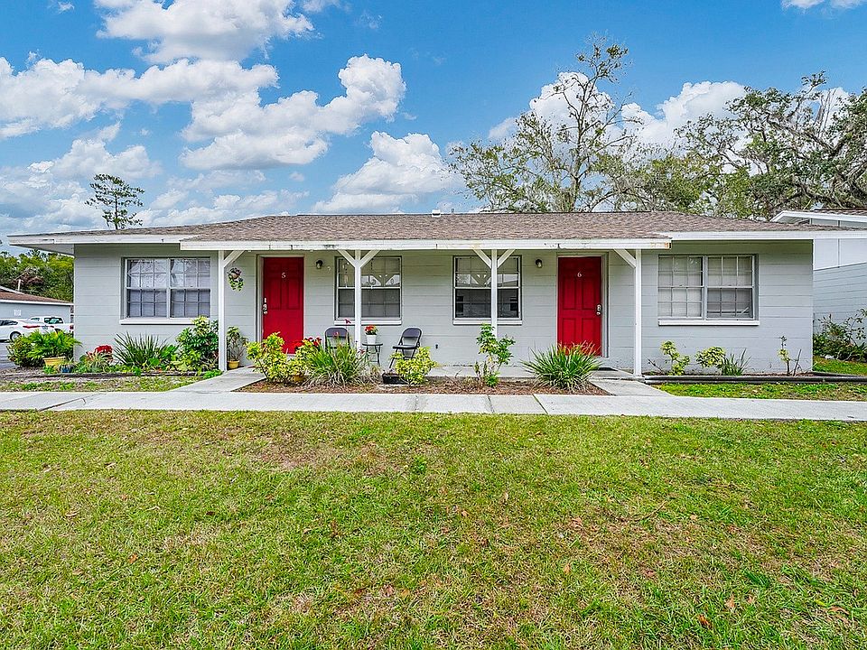 Charming duplex with vibrant red doors, surrounded by lush greenery and a well-maintained front lawn under a bright blue sky.