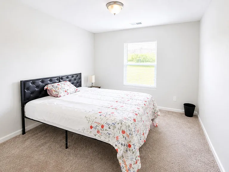 Carpeted bedroom with a full bed, side table, and a large window for natural light.