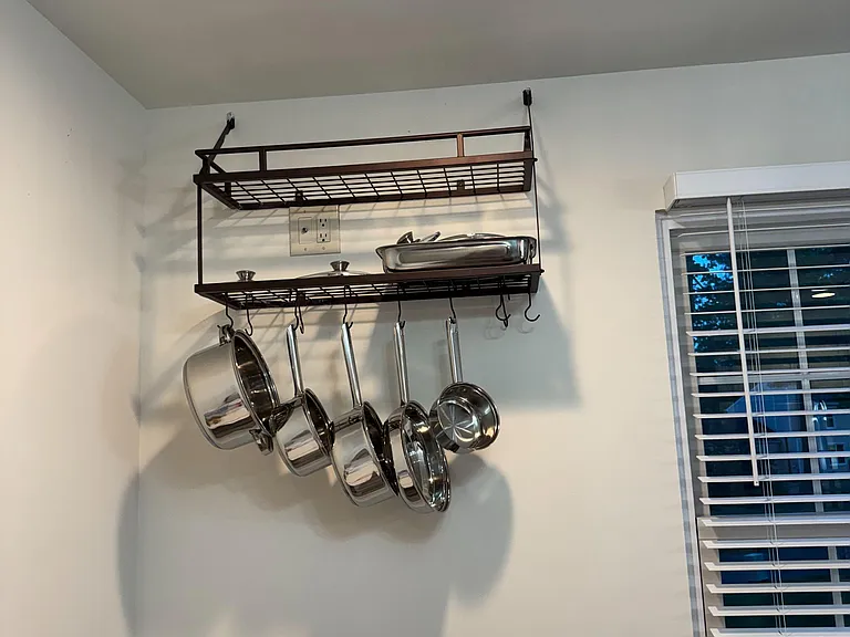 Kitchen includes wall-mounted pot rack with hanging cookware and a nearby window with blinds for added light.