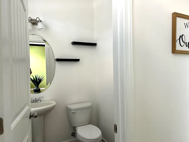 Half bath with pedestal sink, round mirror, wall shelving, and chrome vanity lighting for everyday convenience.