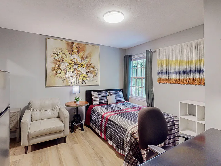 Furnished bedroom with a full bed, desk space, and armchair, plus a large window with curtains and storage shelving.