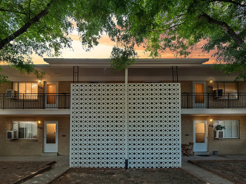 Charming apartment exterior with a cozy brick facade, elegant balcony railings, and ambient evening lighting under a serene sunset sky, nestled among lush green trees.