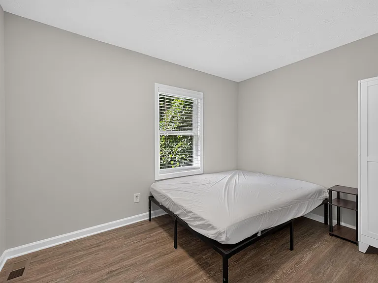Simple bedroom with a bed frame, side table, and a window with blinds, plus a wardrobe for convenient storage.