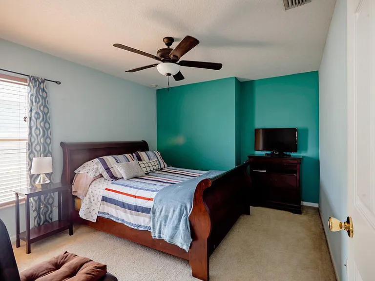 Furnished bedroom with a full bed, ceiling fan, and window with blinds and curtains, plus a TV and dresser for storage.