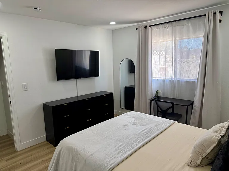Bedroom with a full bed, dresser, wall-mounted TV, and a dedicated desk by the window with full-length mirror.