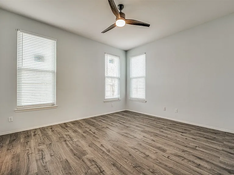 Spacious room featuring wood-finish flooring, light gray wall paint, two windows with blinds, and a ceiling fan with integrated lighting