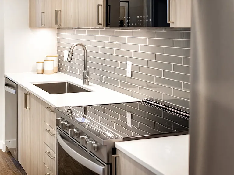 Upgrade Collection I kitchen with light wood cabinetry, grey tile backsplash, white quartz countertops, and stainless steel appliances