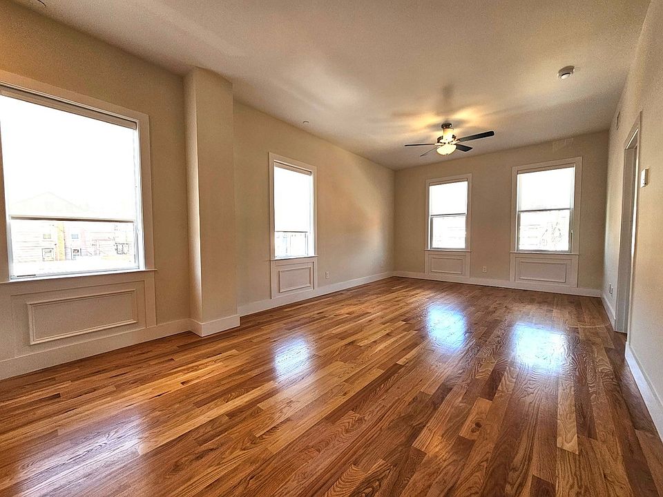 Bright, airy living room featuring gleaming hardwood floors, neutral walls, & abundant natural light from multiple large windows. The spacious layout & ceiling fan create a comfortable, inviting space perfect for relaxing or entertaining.